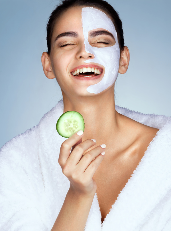 Laughing woman with the slice of cucumber in hand. Photo of funny girl with moisturizing facial mask. Beauty &amp; Skin care concept